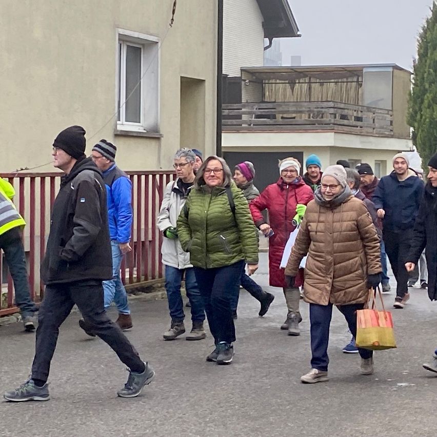 Eine Gruppe von Menschen in Winterkleidung geht gemeinsam auf einer Straße, einige tragen Taschen. Im Hintergrund sind Gebäude mit Balkonen zu sehen.