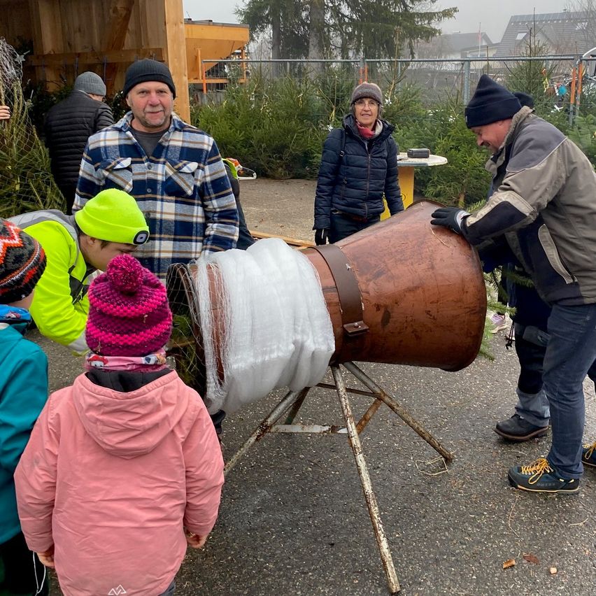 Eine Gruppe von Menschen, einschließlich Erwachsener und Kindern, ist um einen großen, braunen, mit Plastik bedeckten Fass versammelt. Ein Mann justiert das Fass, während ein anderer zusieht. Die Szene spielt sich draußen ab, möglicherweise auf einem Weihnachtsbaumbauernhof.
