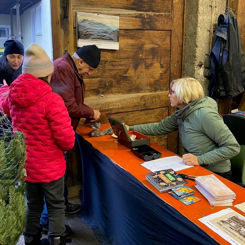 In einem rustikalen Raum übergibt eine Frau einem älteren Mann und einem Kind ein Geschenk. Ein vor ihnen stehender Tisch ist mit blauem und orangefarbenem Tuch bedeckt und hält verschiedene Gegenstände, einschließlich eines Kalenders. Dahinter sind eine Holztür und eine Wand mit einem hängenden Mantel zu sehen.