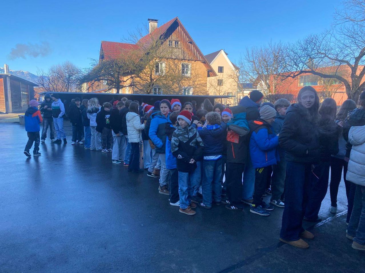 Eine Gruppe von Kindern in Winterkleidung und Mützen steht an einem sonnigen Tag in einer Reihe vor einem Gebäude.