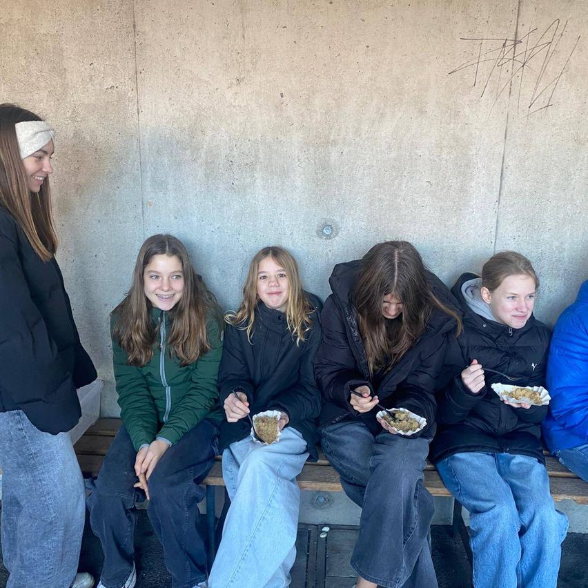Fünf Mädchen sitzen auf einer Bank und essen. Das Mädchen auf der linken Seite lächelt. Vier von ihnen halten Schüsseln mit Essen.