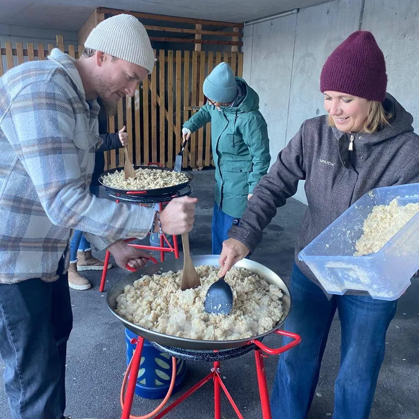 Drei Personen bereiten in großen Pfannen Essen zu, wobei eine Frau einen Behälter mit Zutaten hält. Der Ort ist im Freien mit einem Holzzaun im Hintergrund.