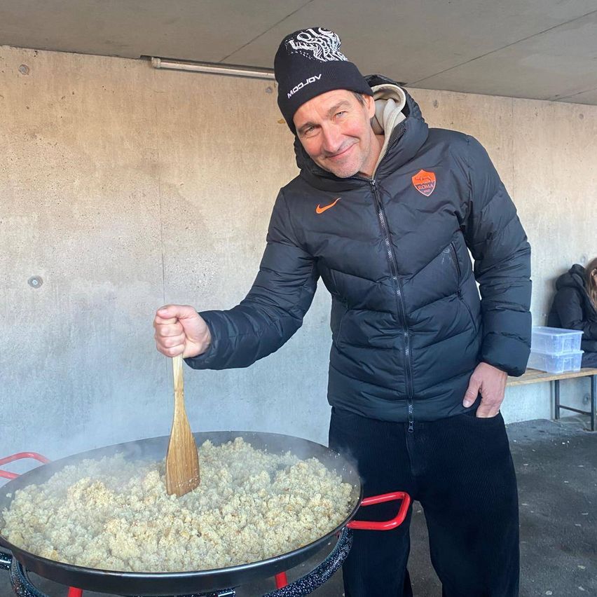 Ein Mann in einer schwarzen Jacke mit einem orangefarbenen Logo auf der linken Brust und einer schwarzen Mütze steht neben einer großen Pfanne, die mit einer Mischung gefüllt ist. Er hält eine Holzspatel und lächelt. Hinter ihm befindet sich eine Betonwand mit einer Bank und einer Aufbewahrungsbox.