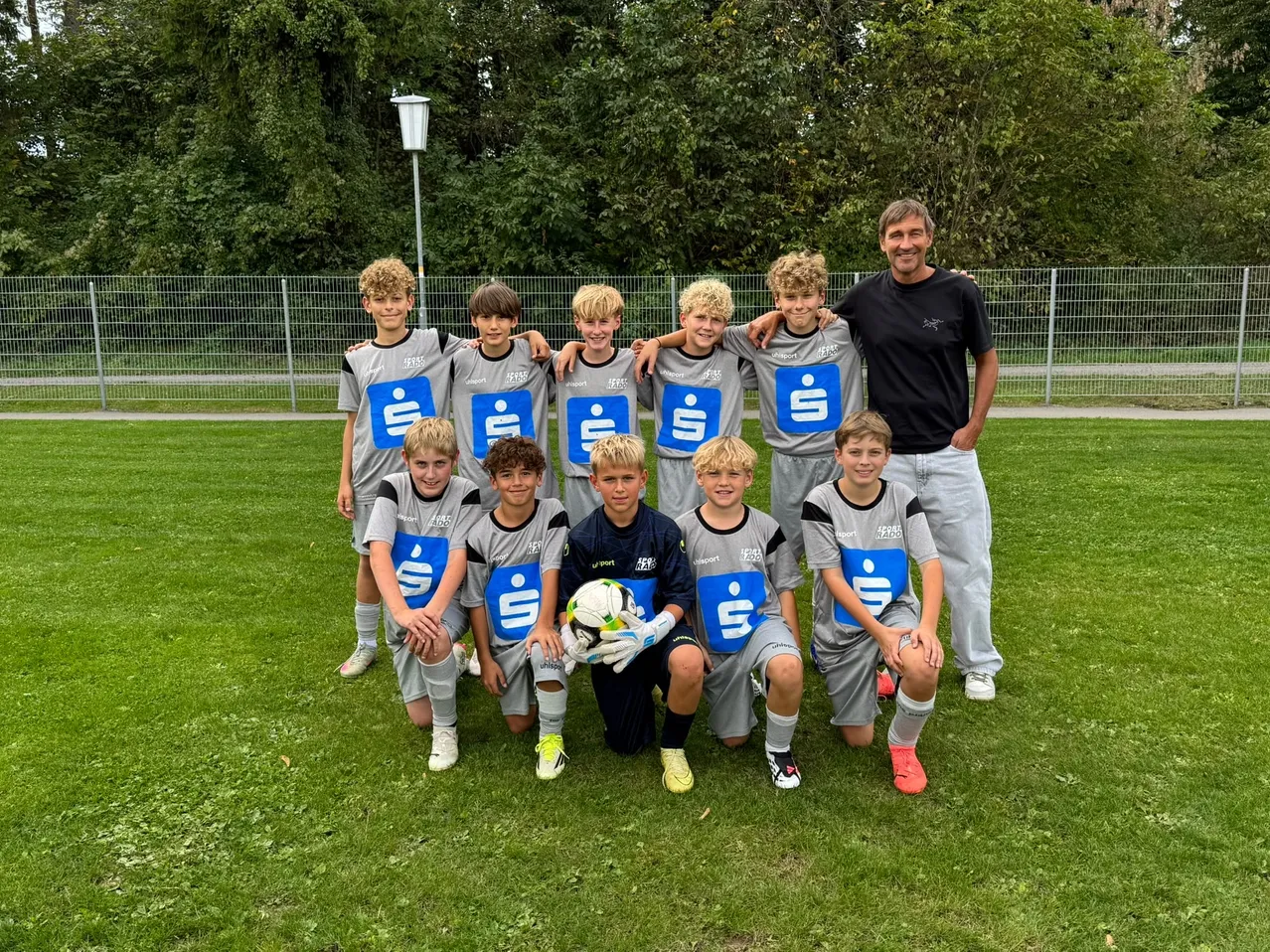 Eine Gruppe junger Fußballspieler, die passende Uniformen mit einem Logo tragen, posiert für ein Foto auf einem grünen Feld.