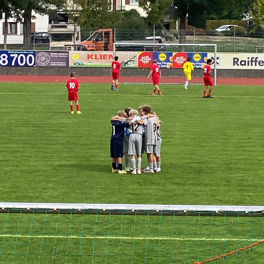 Ein Fußballspiel ist im Gange auf einem Feld mit einem Tor. Die Spieler tragen rote Uniformen und stehen zusammengedrängt.