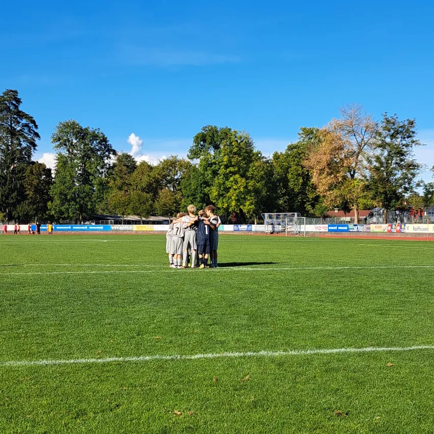 Eine Gruppe junger Fußballspieler steht auf einem Feld versammelt, mit Bäumen und blauem Himmel im Hintergrund.