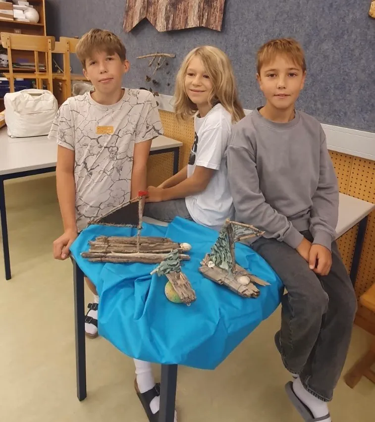Drei Kinder sitzen auf einem Stuhl vor einem Tisch mit einem blauen Tuch. Der Tisch hat ein paar kleine Boote aus Zweigen. Der Junge auf der linken Seite trägt ein weißes Shirt mit einem Design, und der Junge auf der rechten Seite trägt ein graues Langarmshirt. Das Mädchen in der Mitte trägt ein weißes Shirt.