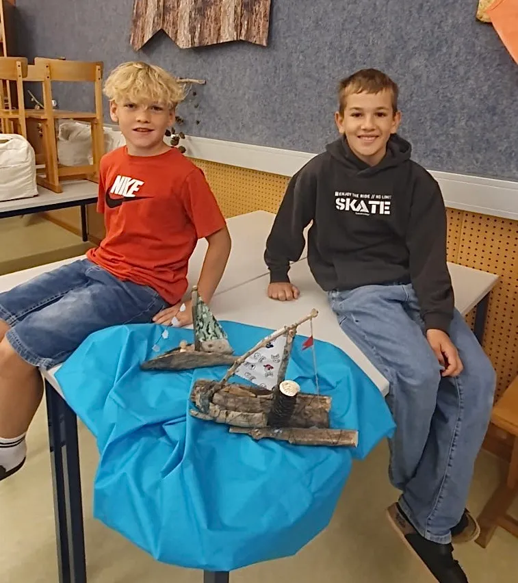 Zwei Jungen sitzen auf einem Stuhl vor einem Tisch. Der Tisch hat ein blaues Tuch und ein kleines Bootsmodell darauf. Der Junge auf der linken Seite trägt ein rotes Shirt mit dem Wort 'NIKE' darauf. Der Junge auf der rechten Seite trägt eine schwarze Kapuzenjacke mit dem Wort 'SKATE' darauf. Hinter ihnen ist eine Wand mit einer hölzernen Dekoration.