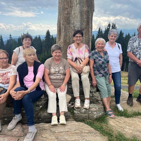 Eine Gruppe älterer Frauen und ein Mann posieren für ein Foto auf einem felsigen Hügel mit einem großen Baumstamm im Hintergrund.