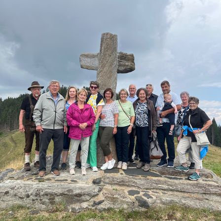 Eine Gruppe von Menschen steht um ein großes Steinkreuz auf einer grasigen Wiese, mit einem bewölkten Himmel im Hintergrund.