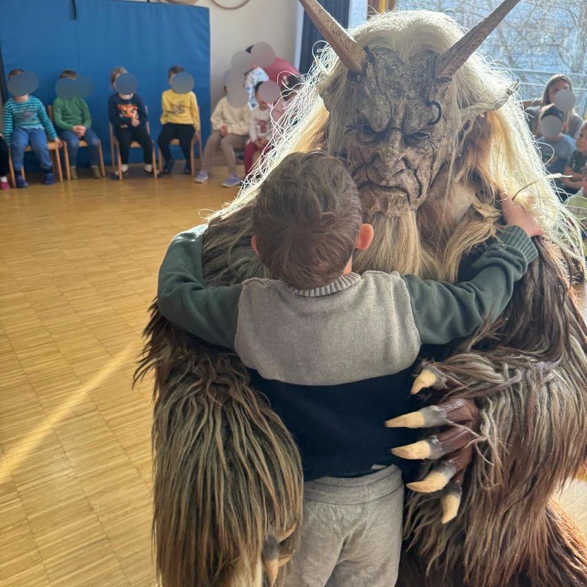 Ein junger Junge steht nah an einer Person in einem gruseligen Monsterkostüm in einer Klassenzimmerumgebung, während mehrere andere Kinder von ihren Plätzen aus zusehen.