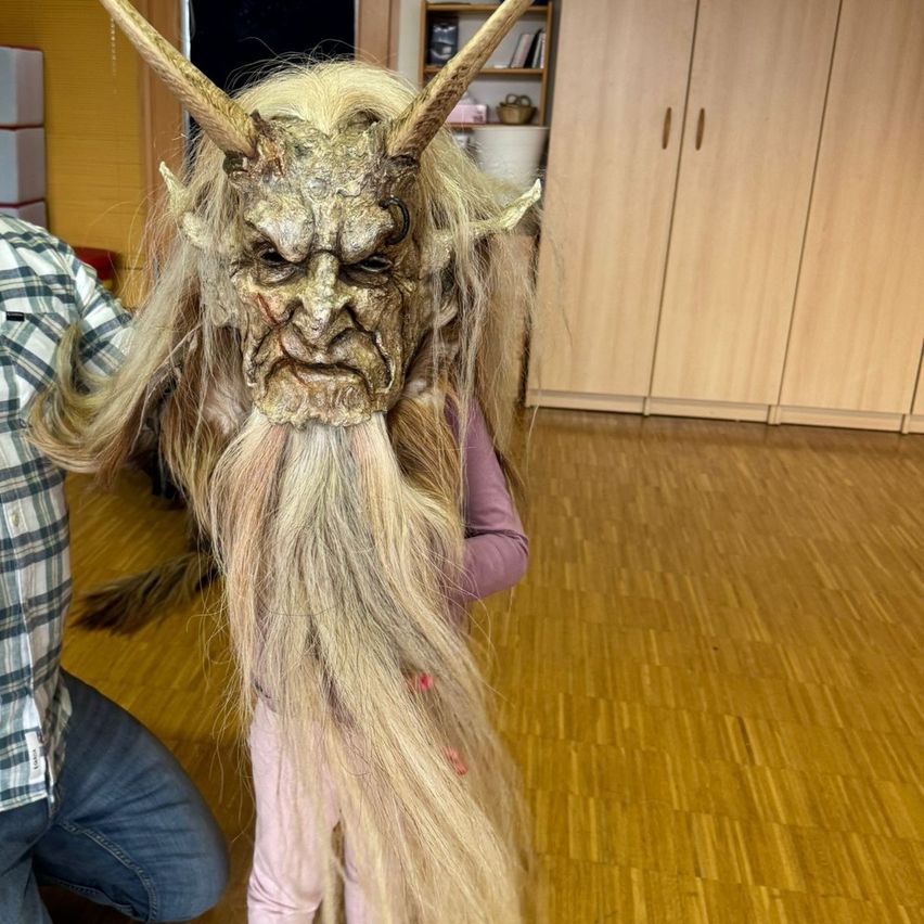 Ein Kind trägt eine detaillierte Dämonenmaske mit Hörnern und langen Haaren in einem Raum mit Holzböden und Schränken.
