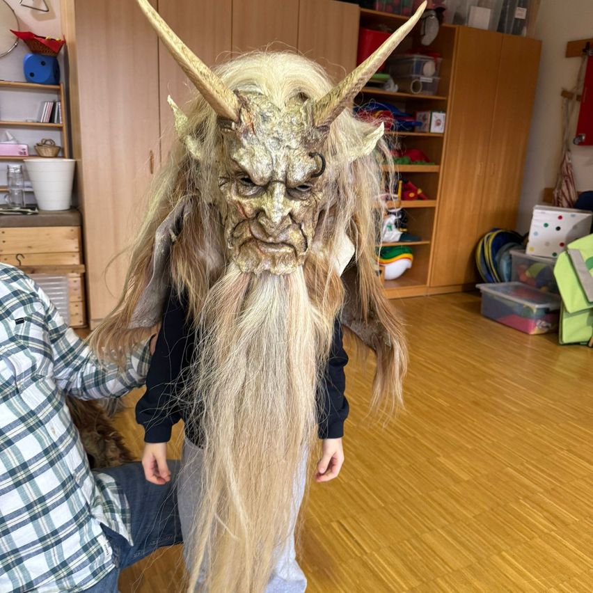 Ein Kind, das eine Hornmaske mit langen blonden Haaren trägt, steht in einem Raum mit Holzböden und Regalen.