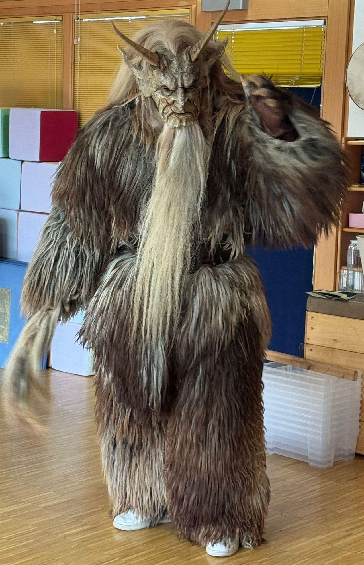 Ein Ganzkörper-Kostüm aus Kunstpelz ist ab der Taille zu sehen. Es hat einen langen Schwanz und einen großen Bart. Das Kostüm steht in einem Raum mit Holzboden.