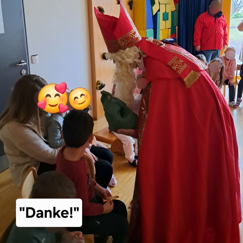 Eine Gruppe von Menschen sitzt auf einem Holzboden, mit einem Mann in Santa-Claus-Kleidung. Lächelnde Kinder erhalten Geschenke. Eine Frau sagt 'Danke!' auf Deutsch und drückt ihre Dankbarkeit aus.