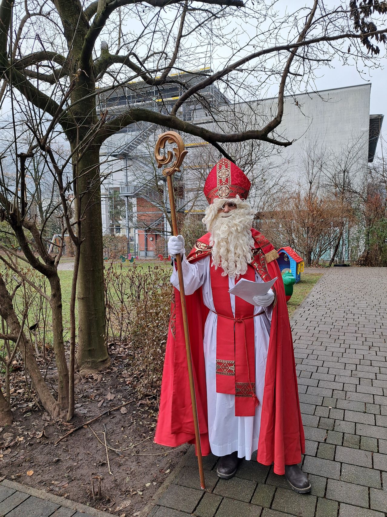 Ein Mann in der Kleidung des Heiligen Nikolaus steht draußen, hält einen Stab und liest ein Papier auf einem Ziegelweg in der Nähe eines Gebäudes.