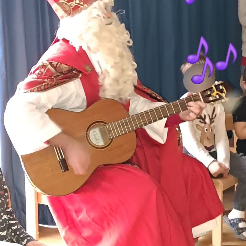 Eine Person in Santa Claus-Kostüm spielt eine Gitarre, während sie auf einem Stuhl sitzt, mit einem Kind vor ihr. Dahinter steht eine Person.
