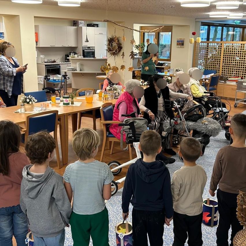 A group of children stand in front of elderly people seated at a table, holding buckets, likely during a festive event. The room has a table with flowers, and several elderly individuals are in wheelchairs. The area is well-lit with decorative lights.