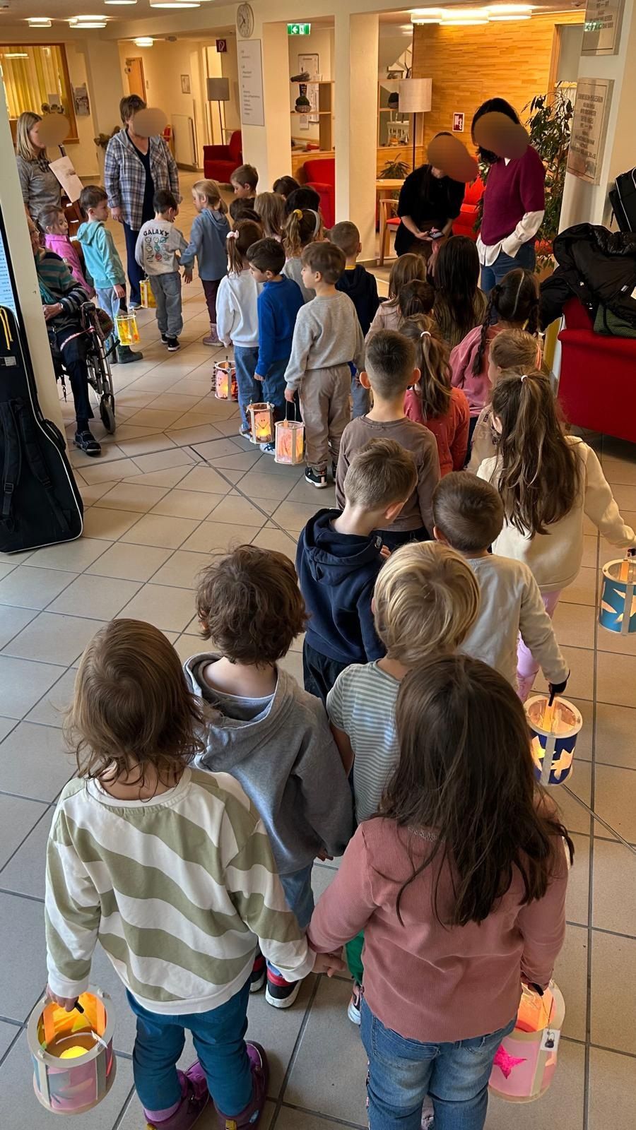 A group of children in a hallway holding candles. Some wear hoodies, while others have long hair. A person in a wheelchair is nearby.