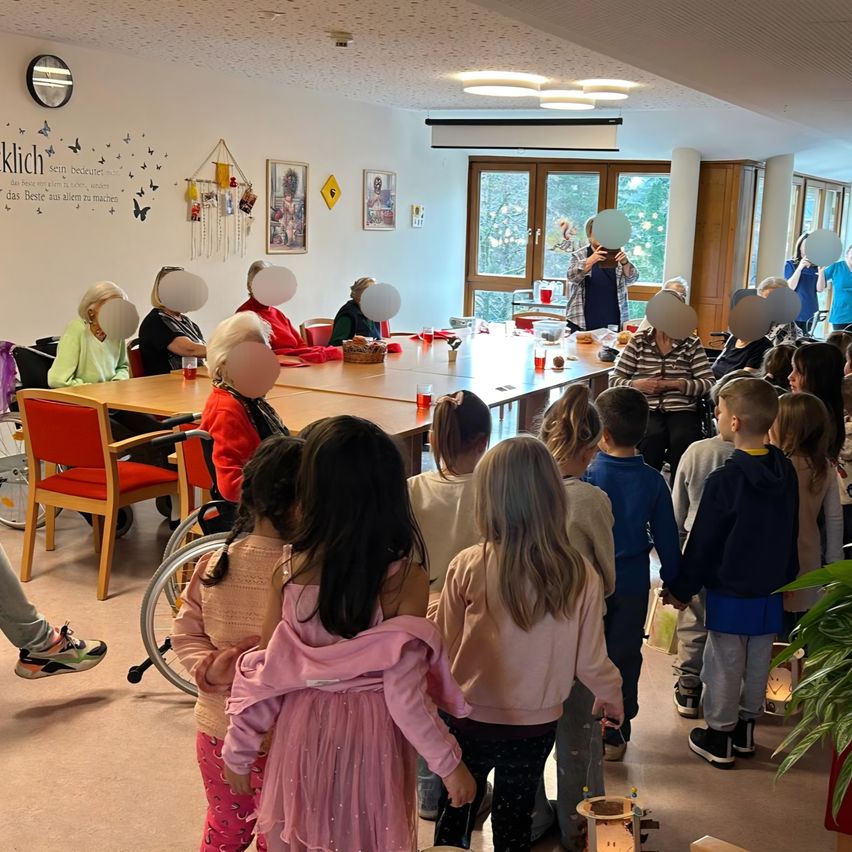 A group of children and adults are in a room. Some adults are seated, while others are standing. One adult is taking a photo. The children are standing in a line. The room has tables, chairs, and decorative items.