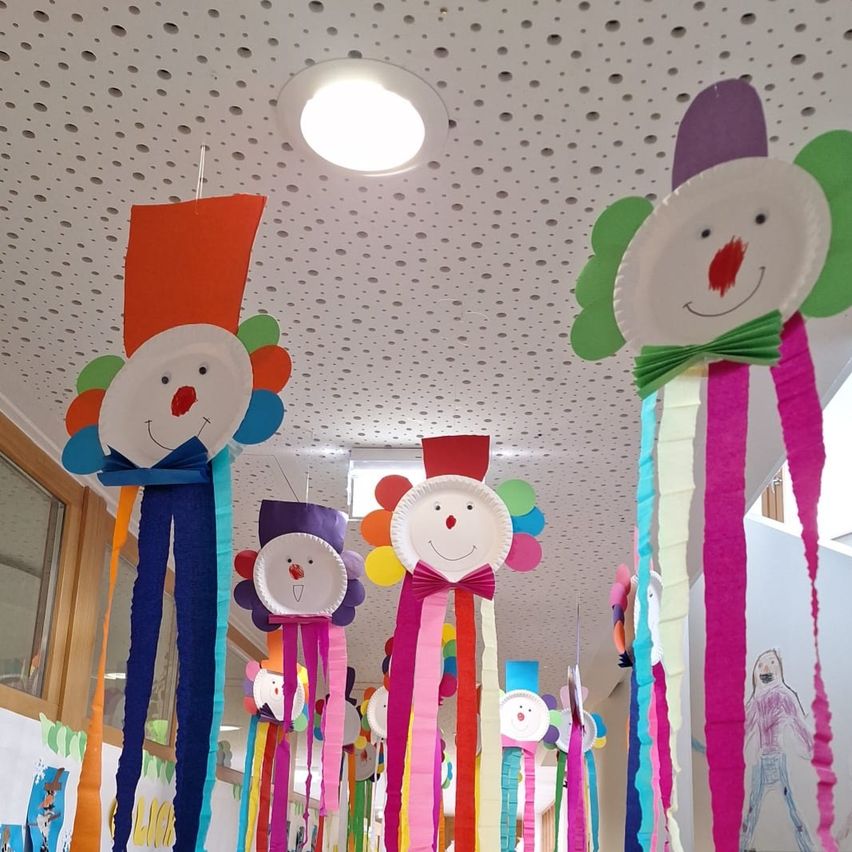 Eine Reihe von farbenfrohen Papierclowns hängt von der Decke, jeder mit roter Nase und Lächeln. Sie sind mit Bändern und buntem Papier dekoriert. Eine helle Lampe ist an der Decke befestigt.