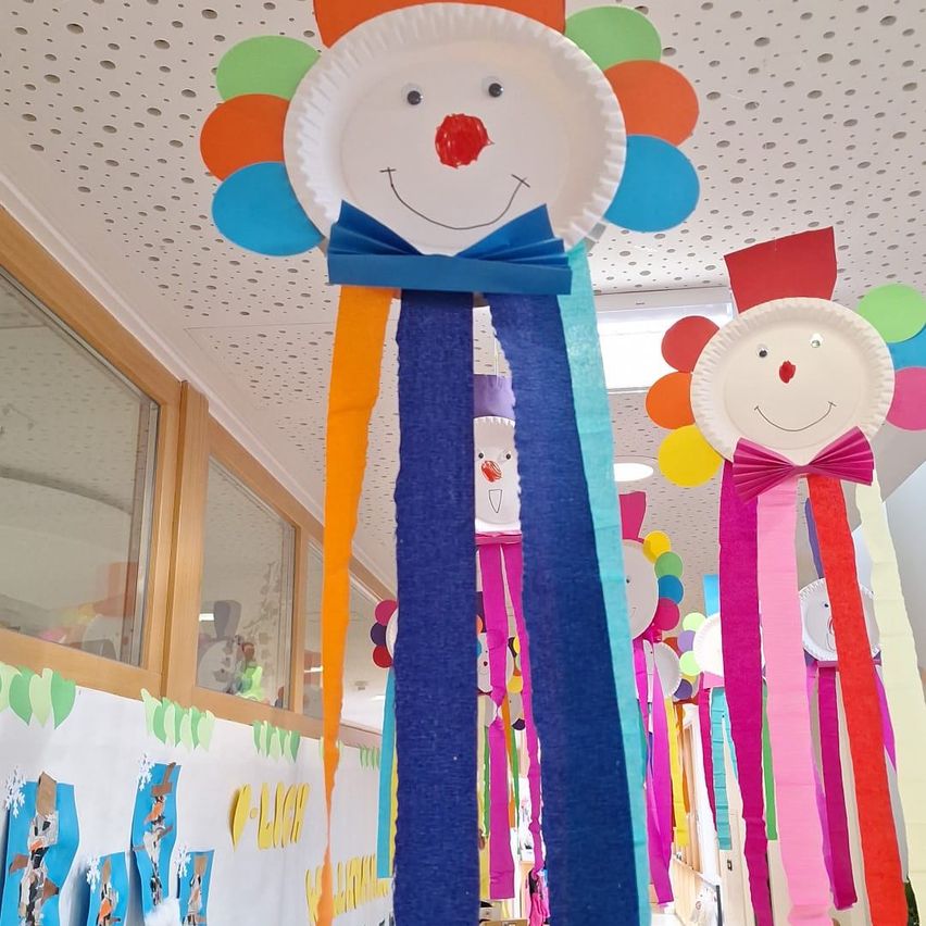Eine fröhliche Papierclown-Dekoration mit bunten Bändern hängt von der Decke, umgeben von anderen ähnlichen Dekorationen und Luftballons.