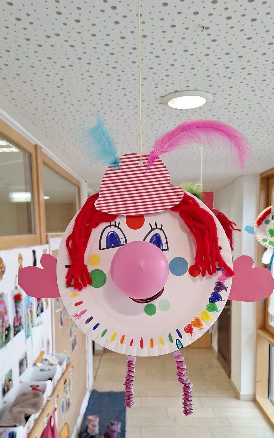 Eine handgefertigte Clown-Dekoration aus Papierteller mit rosa Ballonnase, roter Garnhaar und gestreifter Mütze hängt von der Decke. Sie ist bunt mit verschiedenen Punkten und Herzen.