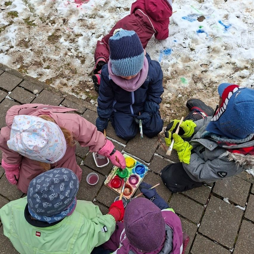 Mehrere Kinder malen auf dem Bürgersteig, umgeben von Schnee. Jedes Kind trägt Winterkleidung und eine Mütze und benutzt Pinsel, um auf einem Malkasten zu malen.