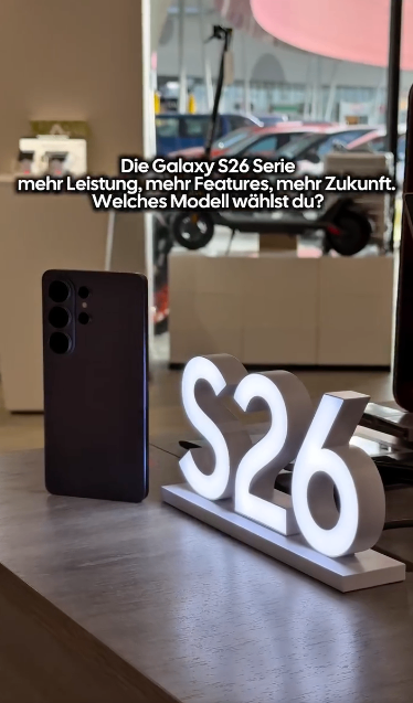 Ein Samsung Galaxy S26 Handy auf einem Holztisch mit einem leuchtenden Schild, das S26 anzeigt. Dahinter ist eine verschwommene Ansicht eines Parkplatzes mit Autos zu sehen.