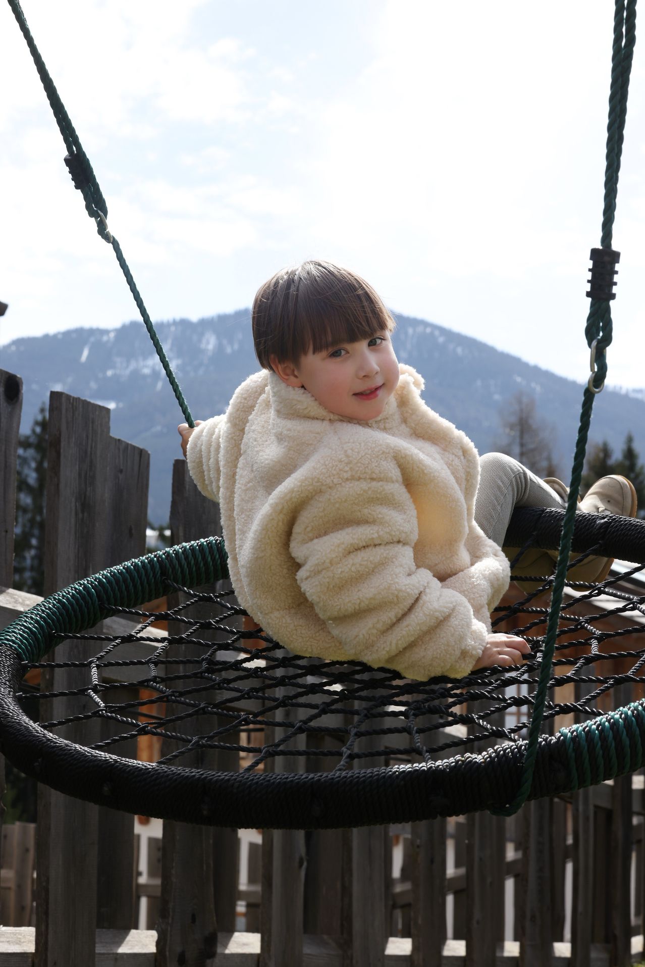Ein Kind sitzt auf einer Schaukel. Die Schaukel ist an einem Holzzaun befestigt. Hinter dem Zaun befinden sich Berge.
