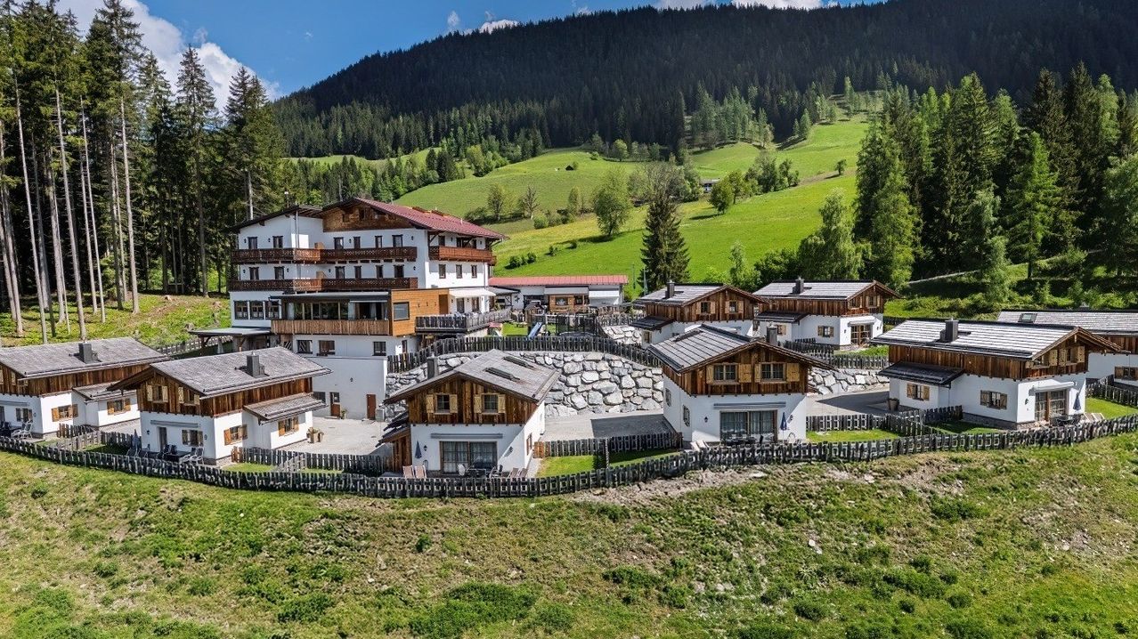 Luftaufnahme eines Hotelkomplexes in den österreichischen Alpen. Die Gebäude sind aus Holz und Stein mit einer Mischung aus braunen und weißen Fassaden. Sie sind von üppigen grünen Wiesen und Bäumen umgeben.
