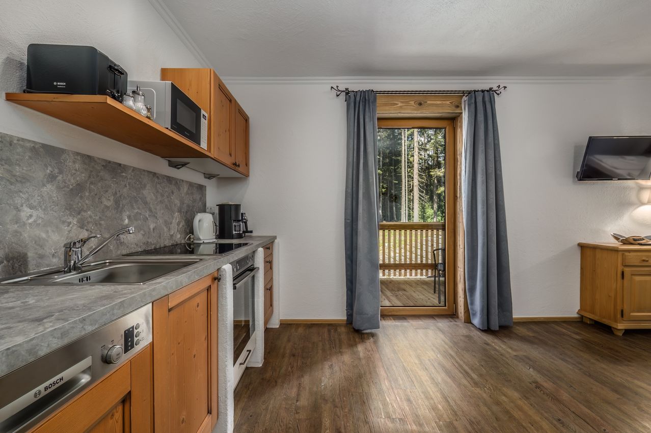 Eine moderne Küche mit Holzschränken und Arbeitsplatten, ausgestattet mit einem Waschbecken, Herd und Mikrowelle. Ein Fenster mit Vorhängen führt zu einer Holzterrasse im Freien.