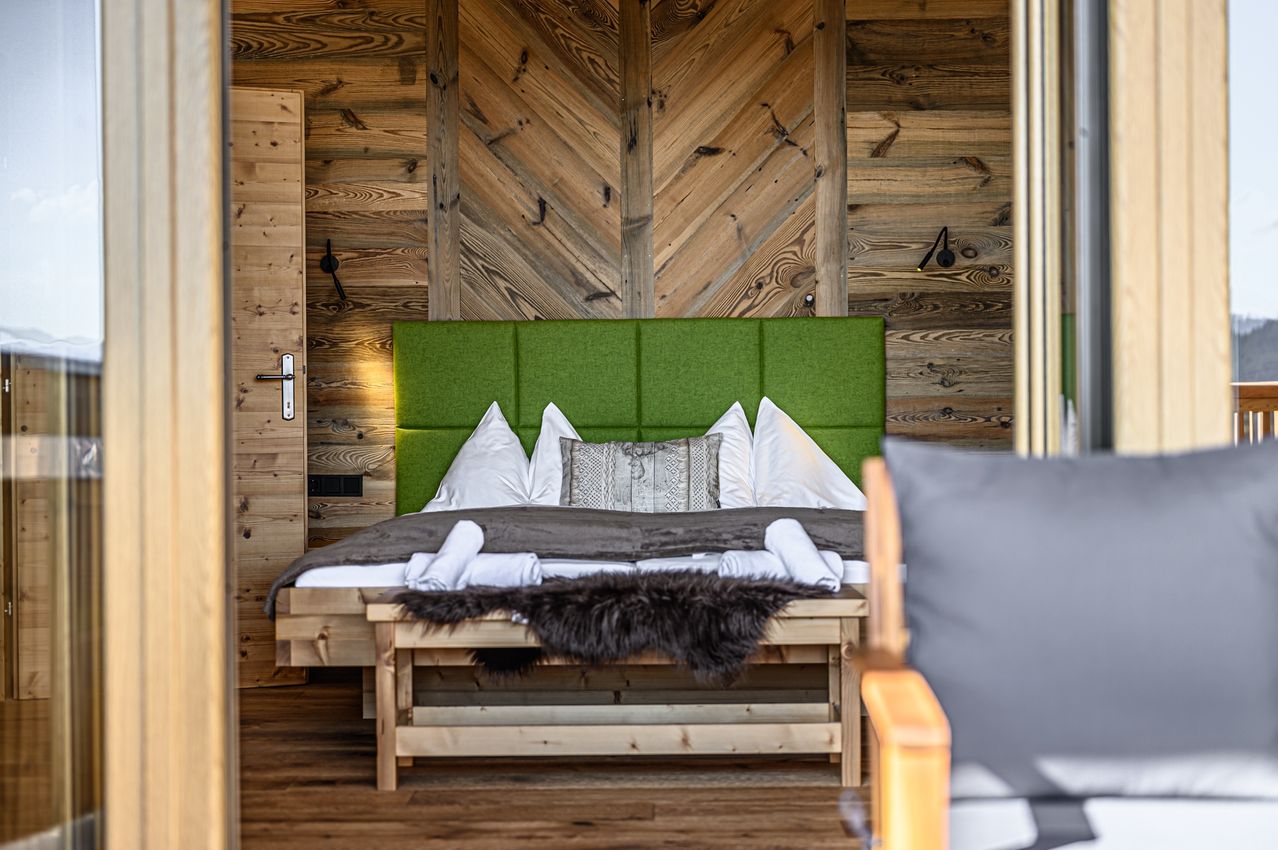 Ein Schlafzimmer mit Holzwänden, einem grünen Kopfteil und einem Bett mit weißen Laken und Kissen. Ein hölzerner Schemel mit Fell und Handtüchern steht vor dem Bett.