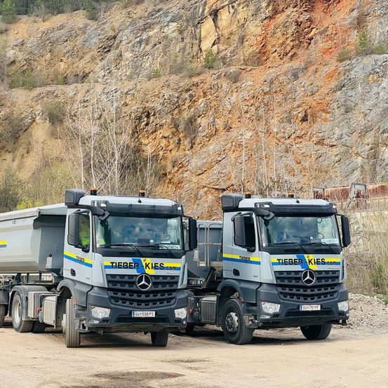 Zwei Mercedes-Benz Trucks mit 'TIEBERKIES.com' auf der Front, geparkt in einem staubigen Bereich mit einem felsigen Fels im Hintergrund.