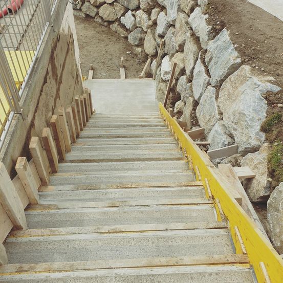 Betonstufen mit gelben Geländern werden vor dem Hintergrund einer Steinmauer errichtet. Die Stufen haben hölzerne Steigungen und werden auf einer Schotteroberfläche gebaut.