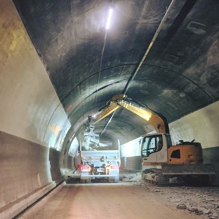 Ein großer Bagger arbeitet in einem Tunnel mit Schutt auf dem Boden und einem Lkw im Hintergrund.