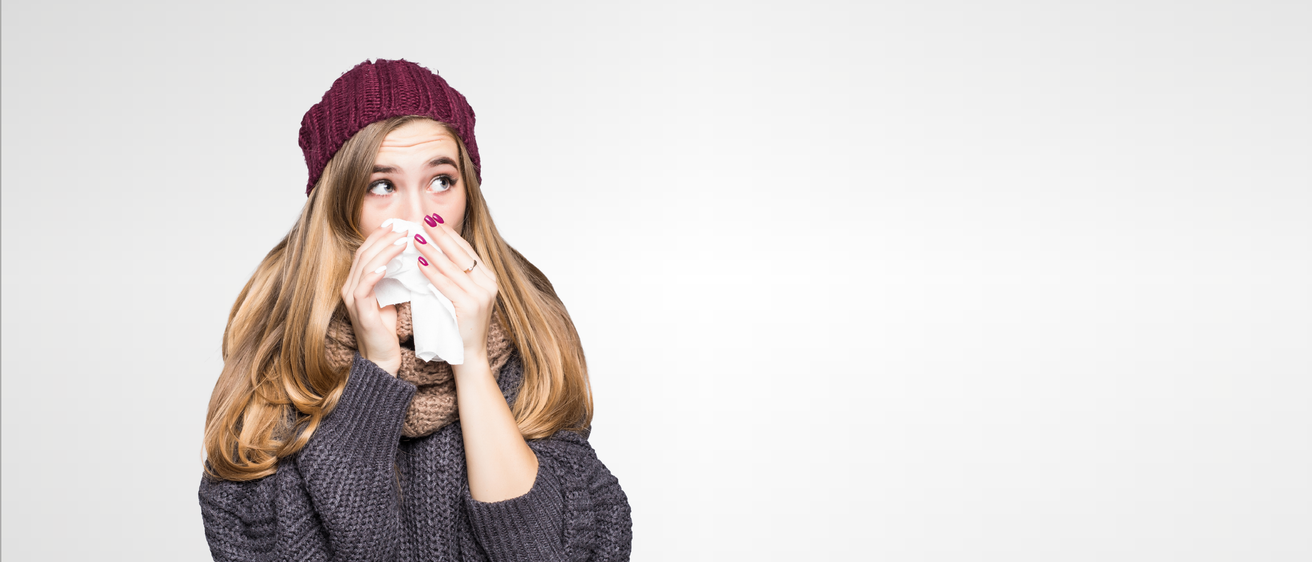 Eine Frau mit einer gestrickten Mütze und einem Schal niest in ein Taschentuch. Sie hat einen Ring am Finger und steht vor einem weißen Hintergrund.
