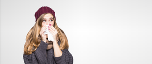 Eine Frau mit einer gestrickten Mütze und einem Schal niest in ein Taschentuch. Sie hat einen Ring am Finger und steht vor einem weißen Hintergrund.