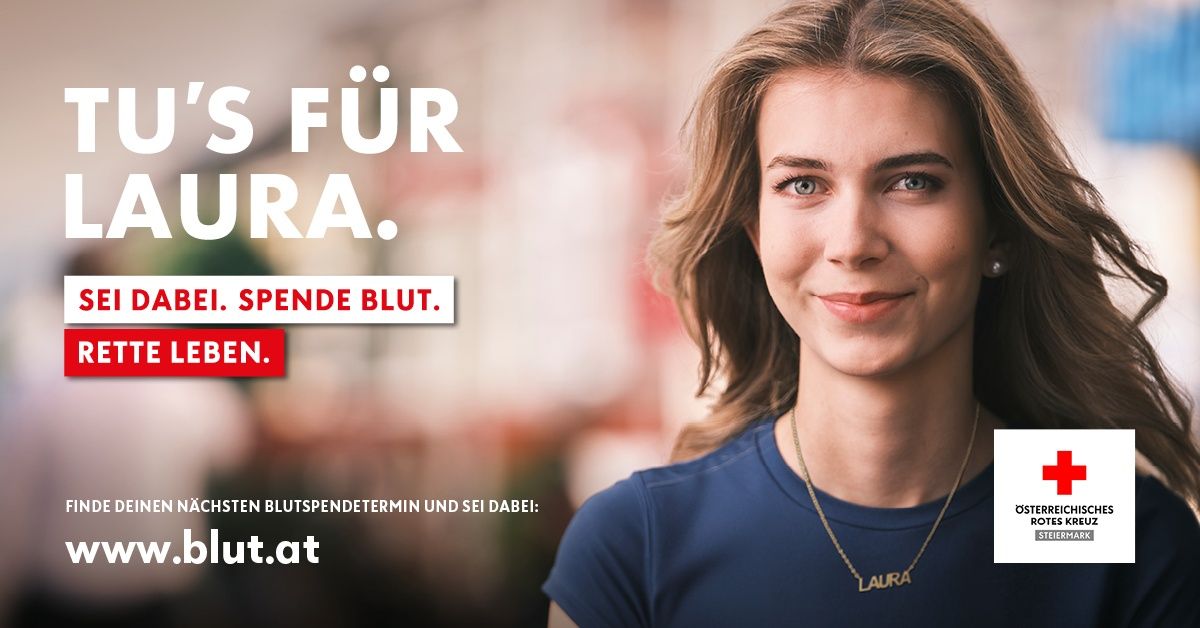 Eine lächelnde Frau mit blonden Haaren trägt ein blaues T-Shirt und eine Halskette mit dem Namen Laura. Der Hintergrund ist verschwommen mit einem roten Banner, das 'Spende Blut' sagt. Das Bild ermutigt zur Blutspende.