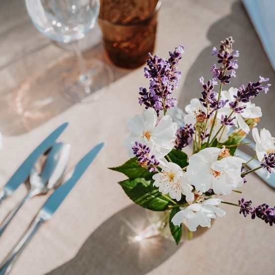 Eine Tischdekoration besteht aus einem Weinglas, Besteck und einer kleinen Vase mit weißen Blumen und lila Lavendel.