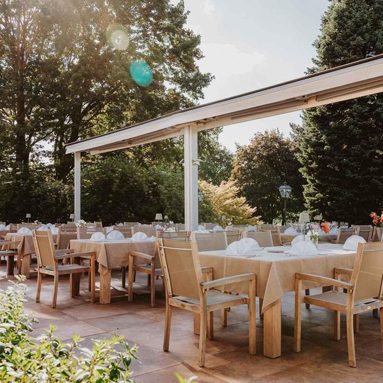 Gartenbereich mit Holztischen und Stühlen, überdacht von einem Vordach, umgeben von Bäumen. Die Tische sind mit weißen Servietten, Blumen und Tellern gedeckt.