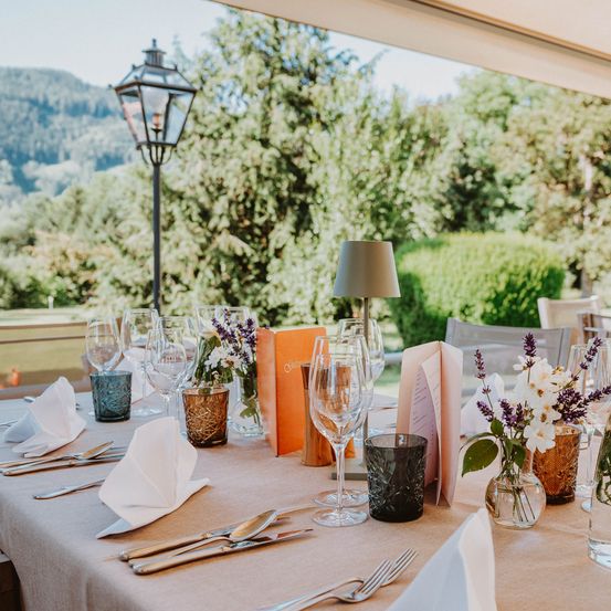 Ein Esstisch im Freien mit Weingläsern, Besteck und Blumen in Vasen. Eine Lampe steht auf dem Tisch und ein Buch liegt darauf. In der Ecke steht ein Laternenpfahl und ein Stuhl steht neben dem Tisch. Der Außenbereich hat eine Bergsicht und es gibt Bäume und Büsche.