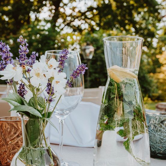 Eine Tischdekoration mit einer durchsichtigen Wasserkaraffe, Glasvasen mit weißen und lavendelfarbenen Blumen, einem Weinglas und einem Kupferbecher auf einer weißen Tischdecke mit verschwommenen Bäumen im Hintergrund.