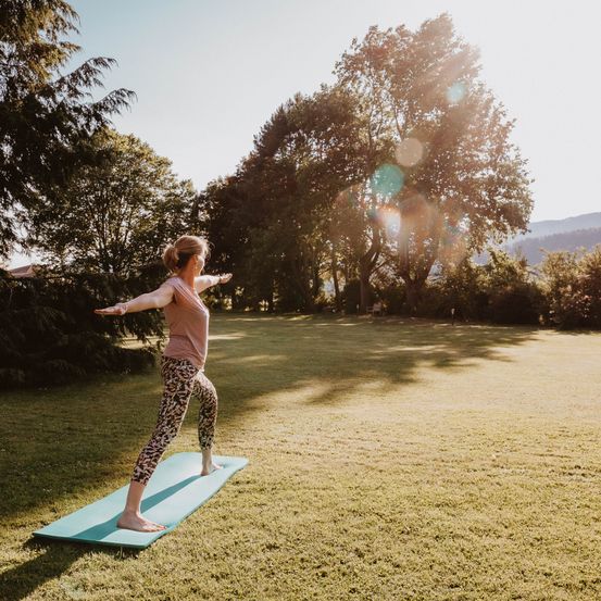 Eine Frau praktiziert Yoga auf einer blauen Matte in einem Grasfeld, umgeben von Bäumen und Bergen. Sie hat die Arme ausgestreckt, und die Sonne scheint hell.