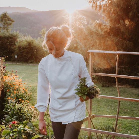 Eine Frau mit Duttfrisur in einer Kochjacke hält einen Bund Kräuter in einem Garten. Die Sonne scheint hinter ihr und beleuchtet den Hintergrund.
