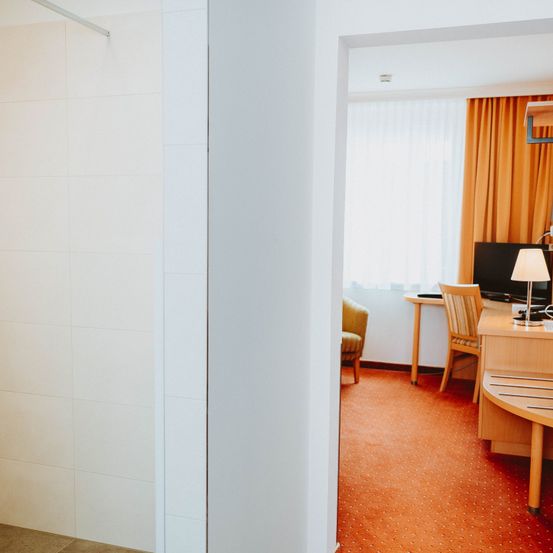 Ein Hotelzimmer mit Schreibtisch, Stuhl und Monitor. Das Zimmer hat eine weiße Wand und einen orangefarbenen Teppich. Es gibt ein Fenster mit Vorhängen.