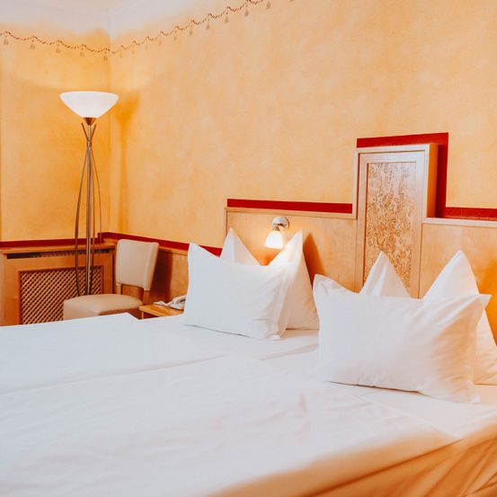 Ein Hotelzimmer mit zwei Betten, jedes mit zwei weißen Kissen, einem hölzernen Kopfteil und einer Lampe. Die Wände sind gelb gestrichen, und es gibt einen Stuhl und einen Heizkörper im Zimmer.