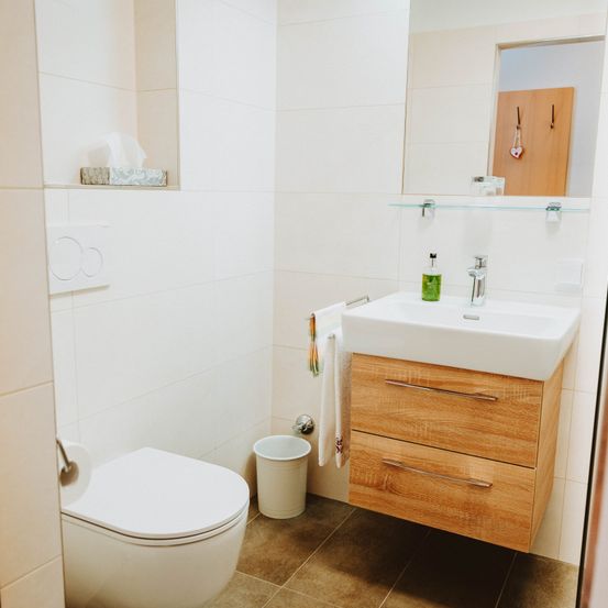 Ein modernes Badezimmer mit weißen Wänden und gefliestem Boden. Es hat eine Toilette, ein Waschbecken mit Wasserhahn und einen Holzschrank mit Schubladen.