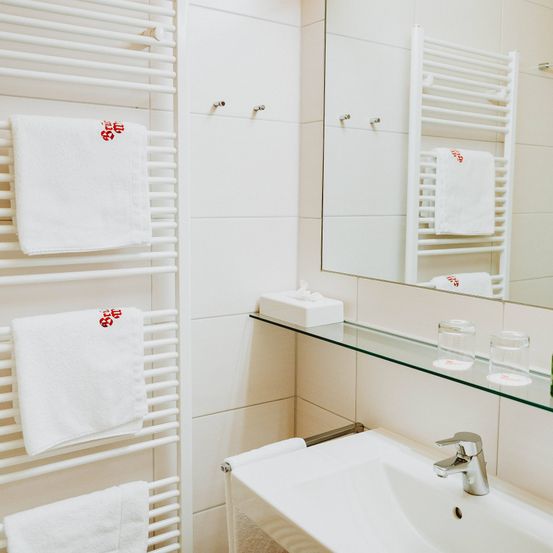 Ein sauberes Badezimmer mit weißen Fliesen und Handtüchern an einem Handtuchhalter. Ein Spiegel reflektiert die Handtücher. Ein Waschbecken mit Wasserhahn und einem Glasregal mit Taschentüchern und Bechern.