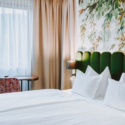 Ein Hotelzimmer mit einem Doppelbett, das mit weißen Laken und Kissen bedeckt ist. Eine Lampe steht auf dem Nachttisch. In der Nähe des Fensters befindet sich ein Schreibtisch mit einem Stuhl.